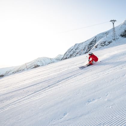 Das Bild zeigt eine Skifahrerin in roter Kleidung, die mit hoher Geschwindigkeit eine frisch präparierte Piste hinunterfährt. Der Schnee wirbelt hinter ihr auf, und im Hintergrund sind hohe Berge zu sehen.