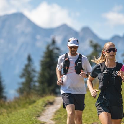 Trailrunnerin und Trailrunner laufen hintereinander, im Hintergrund Bergpanorama