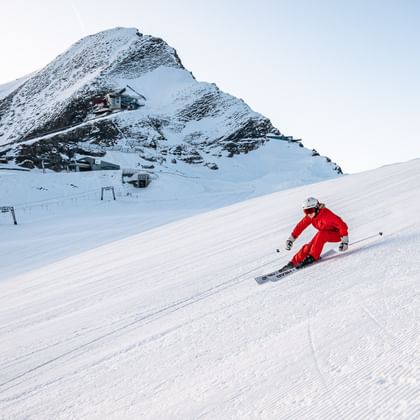 Das Bild zeigt einen Skifahrer in roter Kleidung, der eine schneebedeckte Piste hinunterfährt. Im Hintergrund ist ein felsiger, schneebedeckter Berg mit Gebäuden zu sehen.