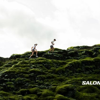 Das Bild zeigt zwei Trailrunner, die über eine mit leuchtend grünem Moos bewachsene Felslandschaft laufen, während der Himmel über ihnen hell und wolkig erscheint; das „Salomon“-Logo ist dezent in der unteren rechten Ecke platziert.
