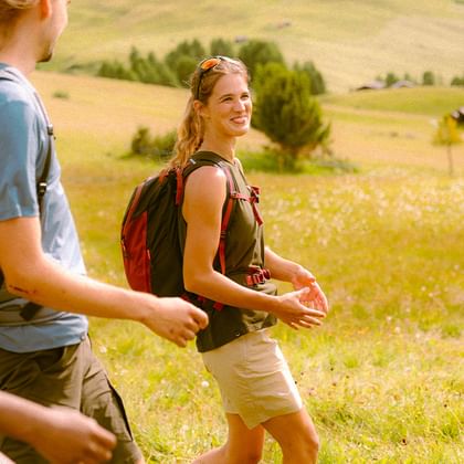 Eine Gruppe junger Wanderer geht lachend durch eine sonnige Almwiese in den Bergen. Sie tragen sportliche Kleidung und Rucksäcke und genießen gemeinsam die Natur.