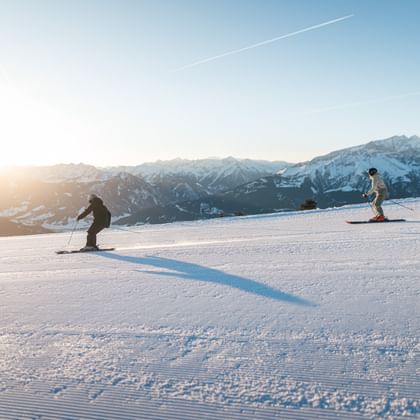 Two skiers descend a groomed ski slope in the low sun. Snow-covered mountains and a blue sky are visible in the background.