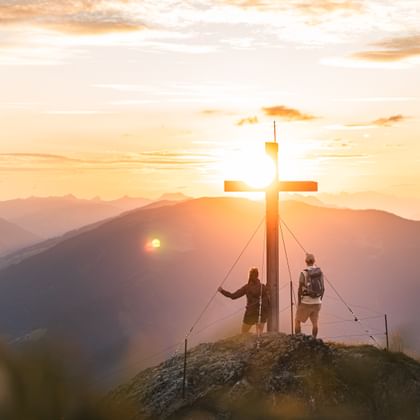 Two people with backpacks stand on a rocky summit beside a large wooden cross, with the sun low on the horizon behind it. The warm sunset light illuminates the surrounding mountains and bathes the valley in golden and orange tones.