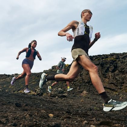 Drei Trailrunner laufen über einen felsigen Bergpfad und tragen Hoka Trailrunning-Schuhe sowie Laufwesten in rauer alpiner Landschaft.