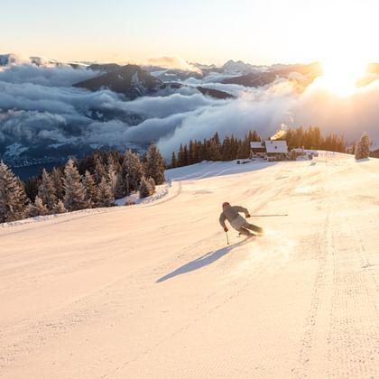 Ein Skifahrer fährt bei Sonnenaufgang auf einer frisch präparierten Piste talwärts, umgeben von verschneiten Bäumen und einer Wolkendecke über dem Tal.