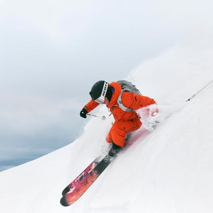 A skier in orange ski gear carves powerfully through fresh powder on a steep slope. Snow sprays up as he makes a dynamic turn.