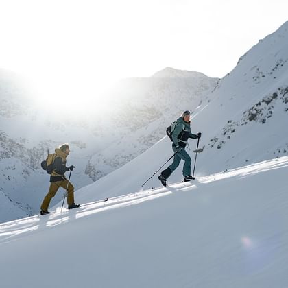 Das Bild zeigt zwei Skifahrer, die mit Tourenski einen verschneiten Berg hinaufsteigen. Die Sonne scheint hinter den Bergen hervor, und die Landschaft ist von schneebedeckten Gipfeln umgeben.