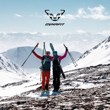 Two ski tourers celebrate their ascent in the snowy Alps, holding touring skis and ski poles, with the DYNAFIT logo visible above them.