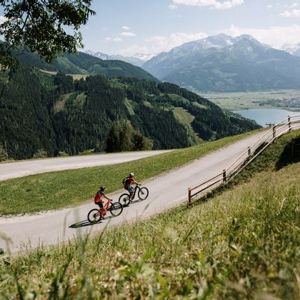 Cyclists ride up the mountain with e-bikes, they are currently in a curve, in the background you can see Lake Zell.
