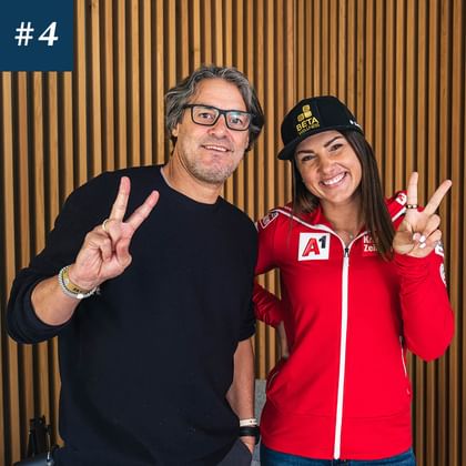 Das Bild zeigt zwei Personen, die beide das Peace-Zeichen machen. Der Mann links trägt eine schwarze Brille und ein schwarzes Oberteil, die Frau rechts trägt eine rote Jacke mit Sponsorenlogos und eine schwarze Mütze. Beide lächeln.