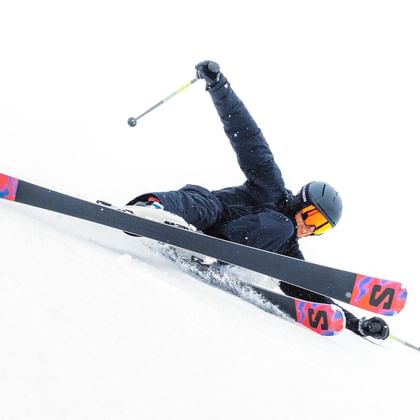 Ein Skifahrer in schwarzer Ausrüstung legt sich dynamisch in die Kurve auf einer schneebedeckten Piste. Der Fahrer trägt einen schwarzen Helm und orange Skibrille, während Schnee unter den Kanten aufspritzt.