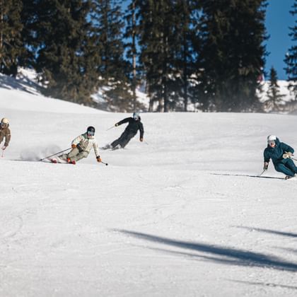 Four skiers make dynamic turns on a snowy ski slope, spraying snow into the air. Tall conifer trees are visible in the background.