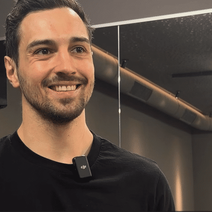 A man with short dark hair and light stubble is smiling and wearing a black T-shirt. A small microphone is attached to his shirt, and mirrors, lights, and a ventilated indoor space can be seen in the background.
