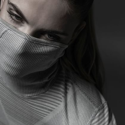 Das Bild zeigt eine Person, die ein eng anliegendes Oberteil trägt, das auch Mund und Nase bedeckt. Die Augen der Person sind intensiv auf die Kamera gerichtet, während das Bild in einem minimalistischen, grauen Farbschema gehalten ist.