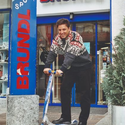 A smiling man stands on a scooter outside a store. “BRÜNDL” appears above the entrance, and “BRÜNDL SPORT” is written on the blue pillar beside it.