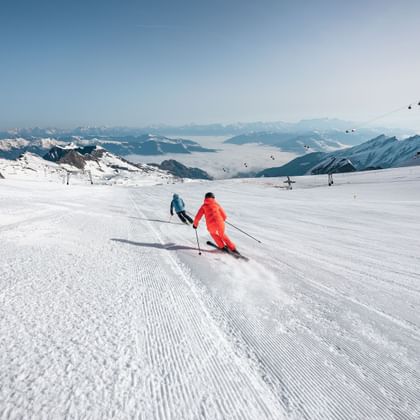 Zwei Skifahrer, einer in blau und der andere in orange, fahren eine breite, frisch präparierte Piste hinunter. Die Berge und die Gondeln im Hintergrund schaffen eine beeindruckende alpine Szenerie.