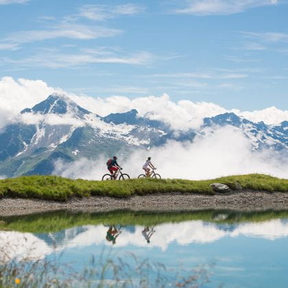 zwei Fahrradfahrer fahren entlang eines Alpensees, worin sich die Berge spiegeln.