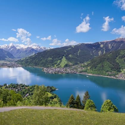 Blick über den Zeller See auf Zell am See, umgeben von grünen Berghängen. Im Hintergrund erhebt sich das schneebedeckte Kitzsteinhorn unter blauem Himmel.