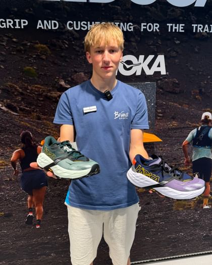 A young man stands in front of a promotional wall, holding a Hoka Speedgoat 7 running shoe in each hand. In the background, there are images of people running and the texts “SPEEDGOAT,” “GRIP AND CUSHIONING FOR THE TRAIL,” and “HOKA” are visible.