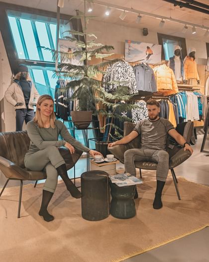Two adults are sitting in armchairs in a sports shop wearing close-fitting functional underwear. In the background, ski clothing, jackets and mannequins can be seen in a modern salesroom. Between the two of them is a small table with cups.