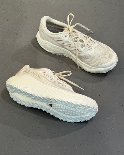 Zwei weiße Sportschuhe liegen auf einem grauen, leicht gesprenkelten Boden. Auf der Seite eines Schuhs ist der Schriftzug „Salomon“ zu lesen, und die Sohlenstruktur ist deutlich sichtbar.