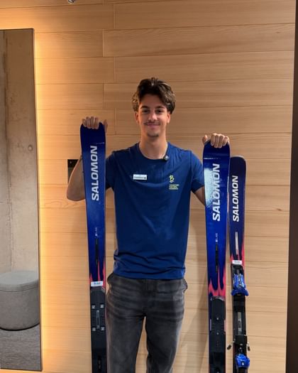 Ein Bründl Sports Mitarbeiter steht vor einer Holzwand und hält zwei blaue Salomon Ski senkrecht in den Händen. Er trägt ein blaues Bründl Sports T-Shirt.