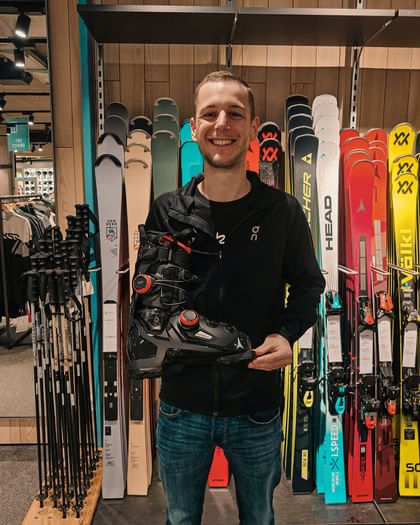 Ein Mitarbeiter in einem Bründl Sports Shop hält den Atomic Hawx Ultra 130 S Dual BOA Skischuh vor einer Wand mit verschiedenen Skimodellen und Stöcken.