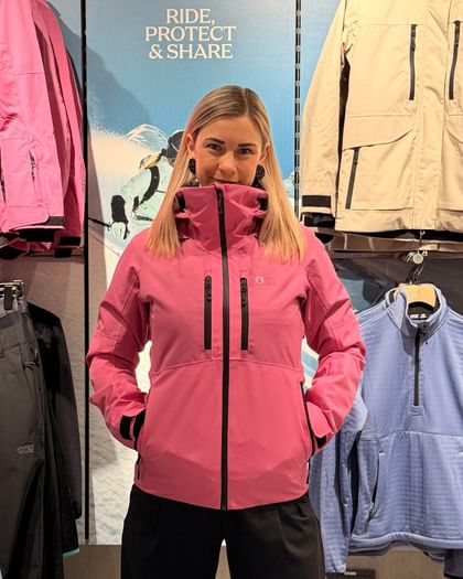 The image shows a person standing front-facing in a sports retail store wearing a pink Picture Sygna jacket. The jacket features a hood, a high collar, a full-length front zipper, and two vertical chest pockets. The hands are placed inside the side zip pockets. In the background, additional ski and snowboard jackets and midlayers are displayed on a Picture-branded wall.