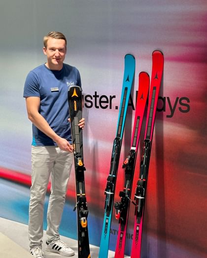 Bründl Mitarbeiter präsentiert neue Atomic Ski Ein Bründl Sports Mitarbeiter steht in einem modernen Showroom vor einer Wand mit dem Atomic-Logo und hält einen schwarzen Ski mit orangefarbenen Details in der Hand. Neben ihm stehen drei weitere Atomic Ski in Blau, Pink und Schwarz.