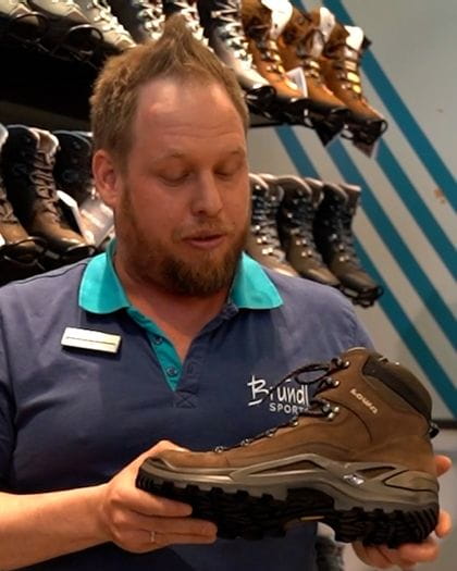 Bründl Sports employee stands in front of a shoe wall and presents the Lowa Renegade hiking shoe.