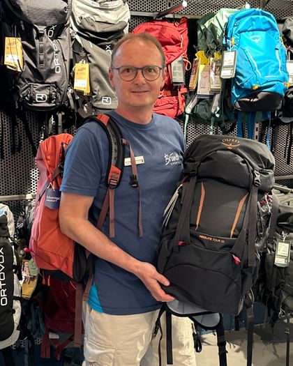 Bründl Sports employee stands with an Osprey Hikelite backpack in front of the backpack wall in the Bründl Sports flagship store