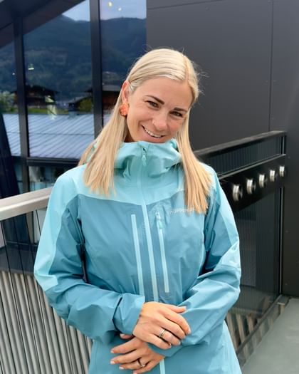 A woman with blonde hair stands on a balcony in front of a modern building with a glass facade, smiling at the camera. She is wearing a light blue Norrona outdoor jacket and pink pants, with mountains and houses visible in the background.
