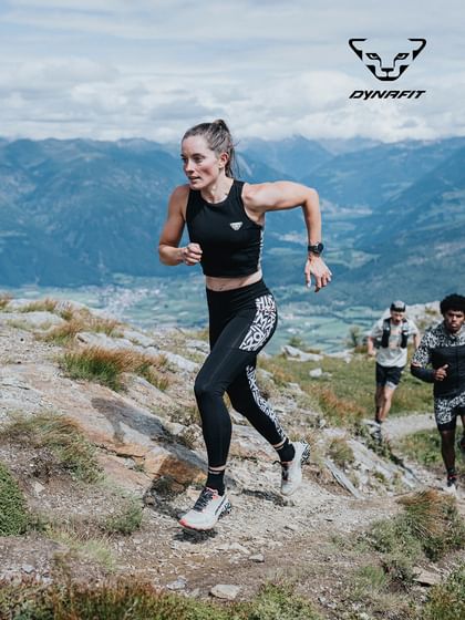 Dynafit trail running athlete running uphill on an alpine mountain trail with panoramic Alps landscape.