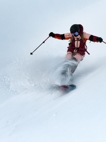 Peak Performance AW 25 A skier glides at high speed through deep powder snow on a steep slope. Snow sprays around as she carves dynamically down the mountain.