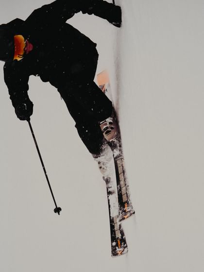 Ein Skifahrer in schwarzer Skibekleidung mit orangefarbener Skibrille ist während eines Sprungs zu sehen. Die schwarzen Atomic Ski, weißen Skischuhe und aufgewirbelten Schneepartikel sind deutlich erkennbar.
