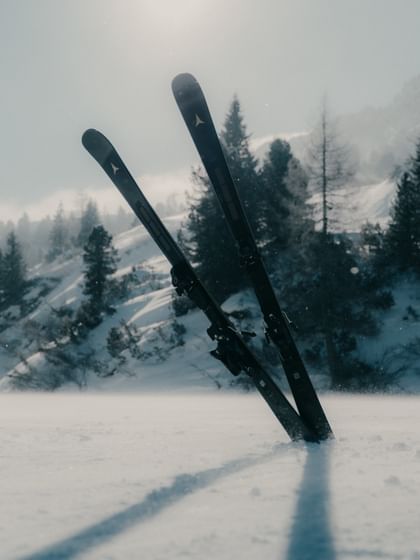 Zwei schwarze Atomic Cloud Ski mit goldfarbenen Logos stecken gekreuzt im Schnee. Im Hintergrund sind verschneite Berghänge und Nadelbäume bei leichtem Schneefall zu sehen.