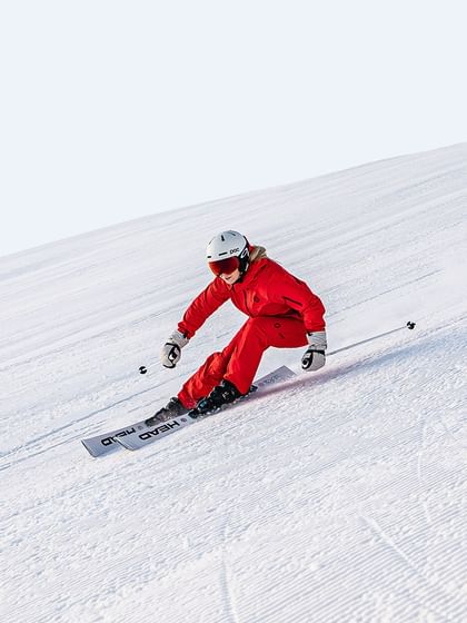 A female skier in a red Scott ski suit carves turns through the snow on a white Head ski. She is also wearing a white POC ski helmet and black Oakley ski goggles.