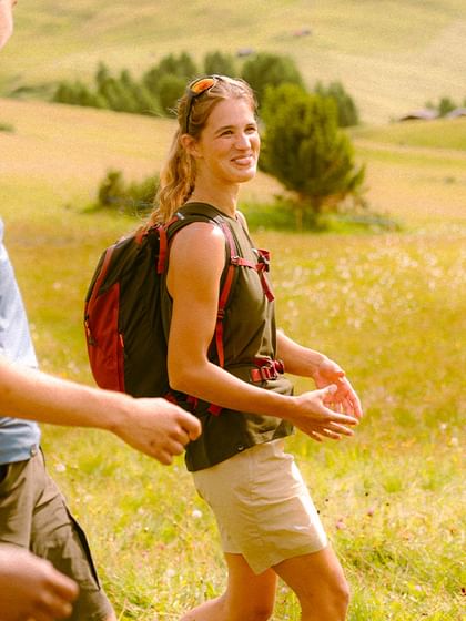 Eine Gruppe junger Wanderer geht lachend durch eine sonnige Almwiese in den Bergen. Sie tragen sportliche Kleidung und Rucksäcke und genießen gemeinsam die Natur.