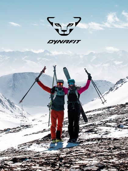 Two ski tourers with DYNAFIT equipment raise their ski poles and carry touring skis in a snow-covered alpine mountain landscape.