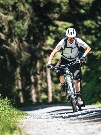 Eine Person mit weißem Helm, Sonnenbrille und Rucksack fährt mit einem Mountainbike einen schmalen Schotterweg durch einen dichten Wald entlang. Sonnenlicht fällt durch die hohen Bäume und beleuchtet Teile des Weges und der grünen Vegetation.