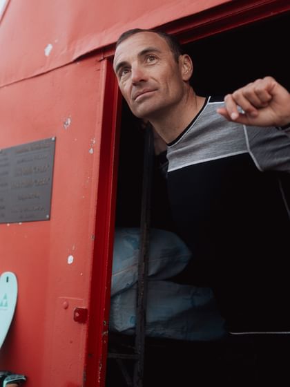 Real moments in the mountains – with Löffler, functional and mindful. A man looks out from a red mountain hut into the snowy alpine landscape. Light blue skis stand next to the door, featuring Löffler apparel.