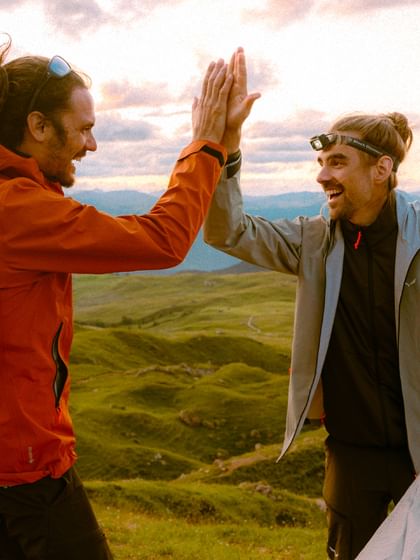 Zwei Männer in Outdoorbekleidung geben sich ein High-Five auf einem Berggipfel bei Sonnenuntergang. Sie lachen, tragen Funktionsjacken und genießen den Moment nach dem Aufbau eines Zelts.