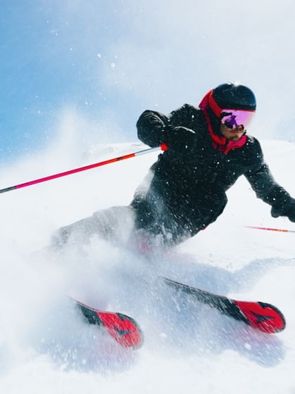 Skifahrer macht eine Kurve im Schnee mit Atomic Redster Ski, Schneespritzer in der Luft, blauer Himmel im Hintergrund.