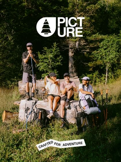 A group consisting of 2 women and 2 men in Picture Organic Clothing outdoor clothing take a break on a large tree trunk in a forest clearing. 