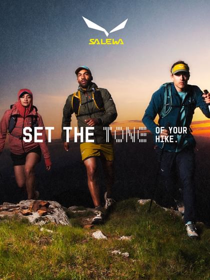 Three people wearing backpacks and outdoor clothing walk along a grassy, rocky mountain path as the sun sets on the horizon. The image includes the text “SET THE TONE OF YOUR HIKE.” and the logo “SALEWA”.