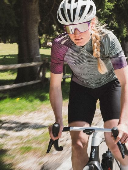 A focused cyclist in athletic clothing rides on a sunny country road, followed by another cyclist.