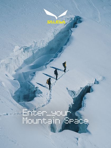 Three mountaineers carrying backpacks walk single file along a narrow snow trail between deep glacier crevasses, photographed from above. The image features the Salewa logo and the text “Enter your Mountain Space”.