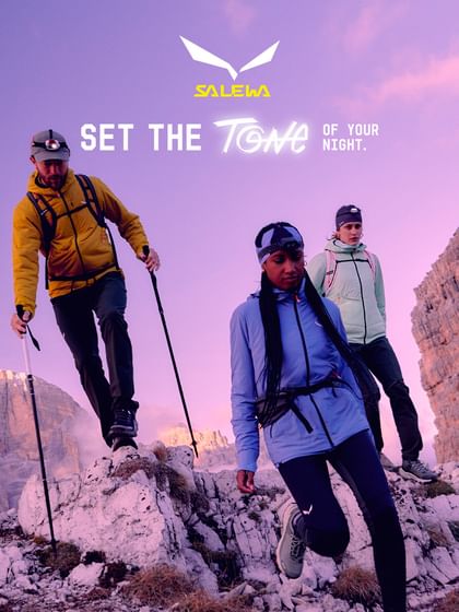 Three hikers with headlamps and trekking poles explore rocky alpine terrain at dusk. They are wearing colorful outdoor gear by Salewa. In the background, the mountains and sky glow in warm purple and pink hues.