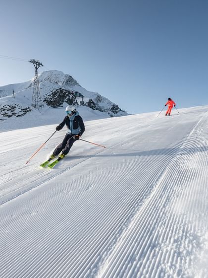 Zwei Skifahrer fahren eine frisch präparierte Piste hinunter, einer in dunkler Kleidung vorne, der andere in leuchtendem Orange dahinter.