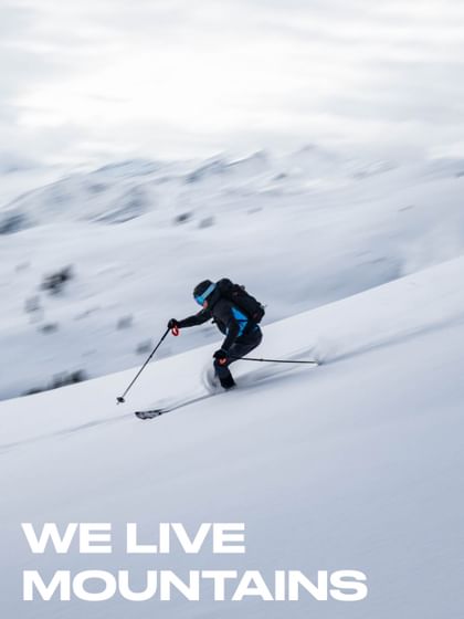 Das Bild zeigt einen Skifahrer, der dynamisch eine schneebedeckte Piste hinunterfährt, umgeben von einer verschneiten Berglandschaft. Unten links steht der Schriftzug „WE LIVE MOUNTAINS“.